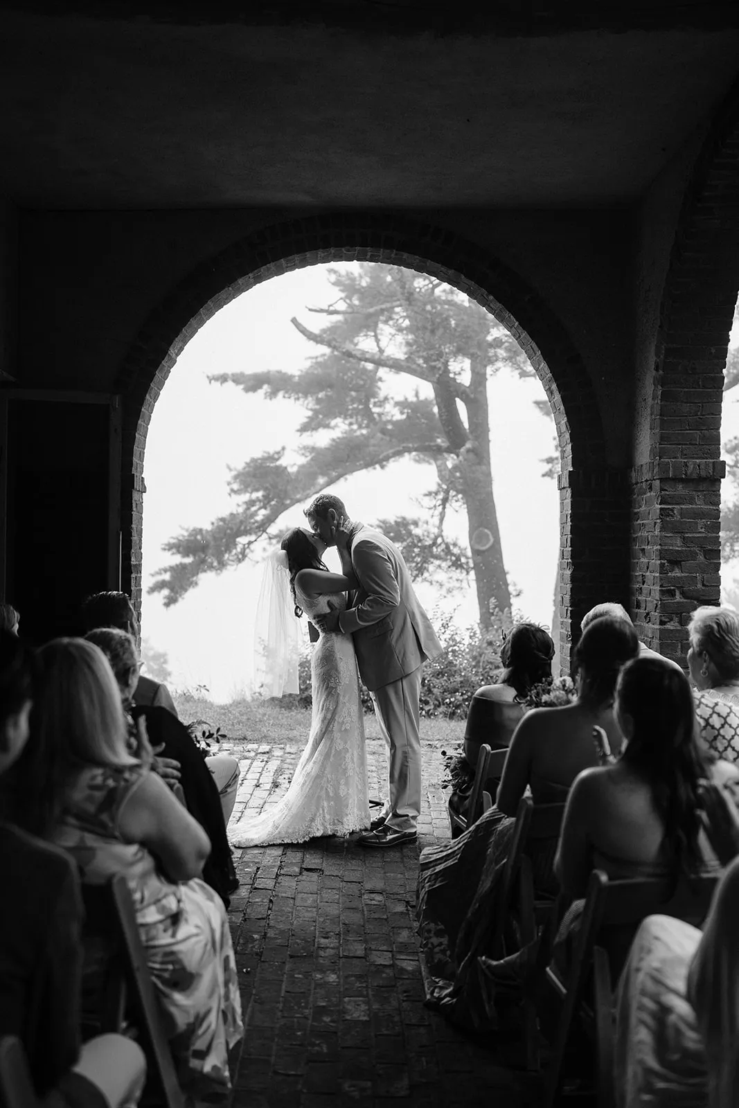 Seated guests watching a couple kiss, framed by the arch with pines and the bay beyond, in black and white