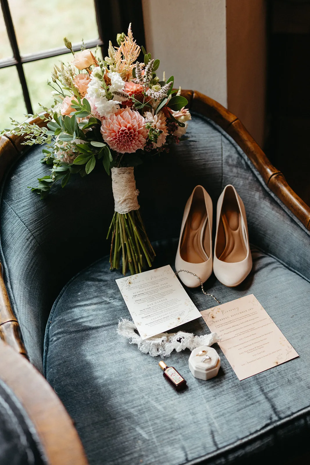 A bouquet of peach dahlias and roses on a velvet chair by the window, with shoes, vows and an invitation