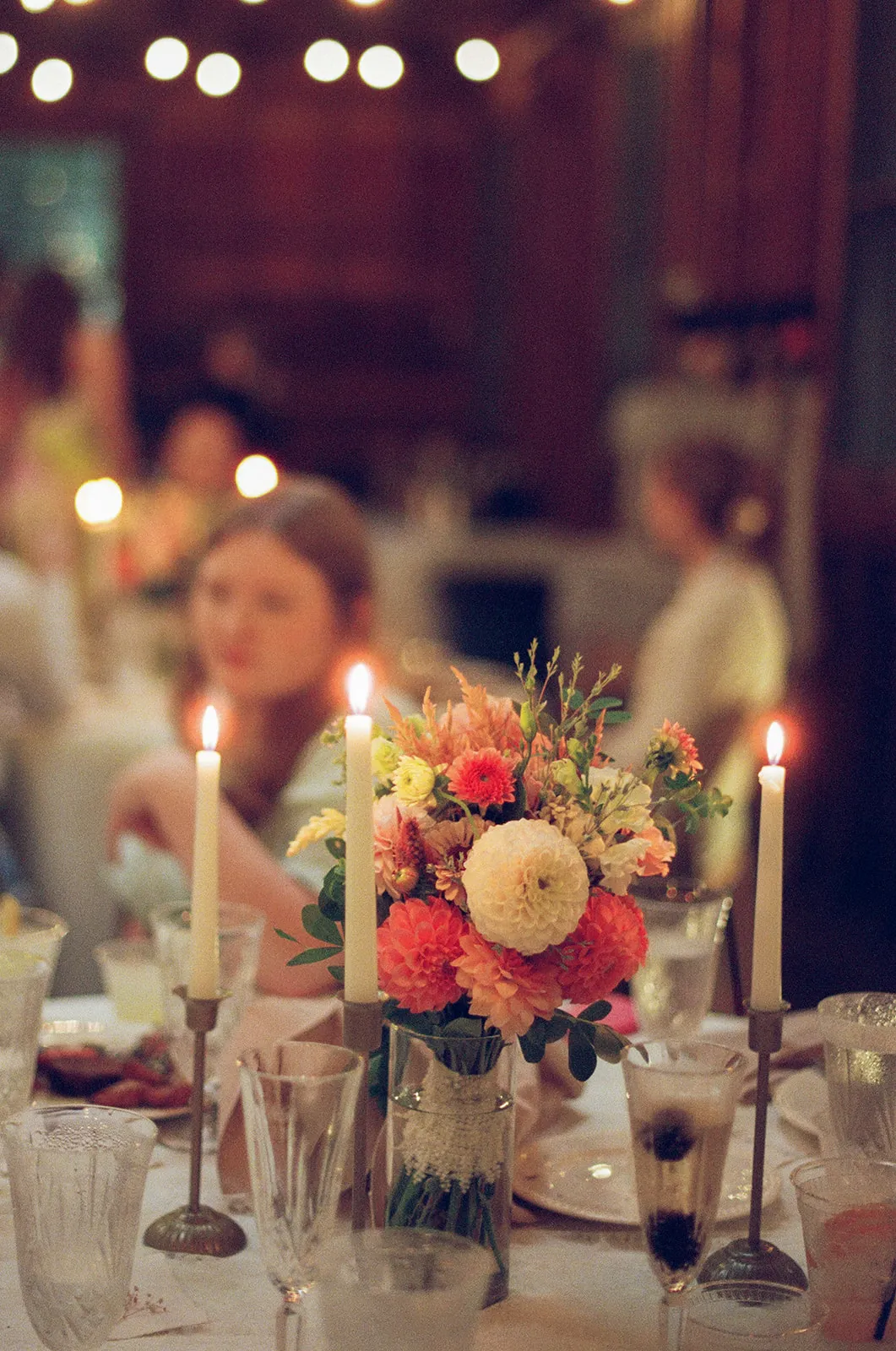 A candlelit dinner table with dahlias, guests talking in the background